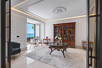 Elegant white room with ornate ceiling, classic furniture, and a balcony offering stunning views of the blue sea and sky.