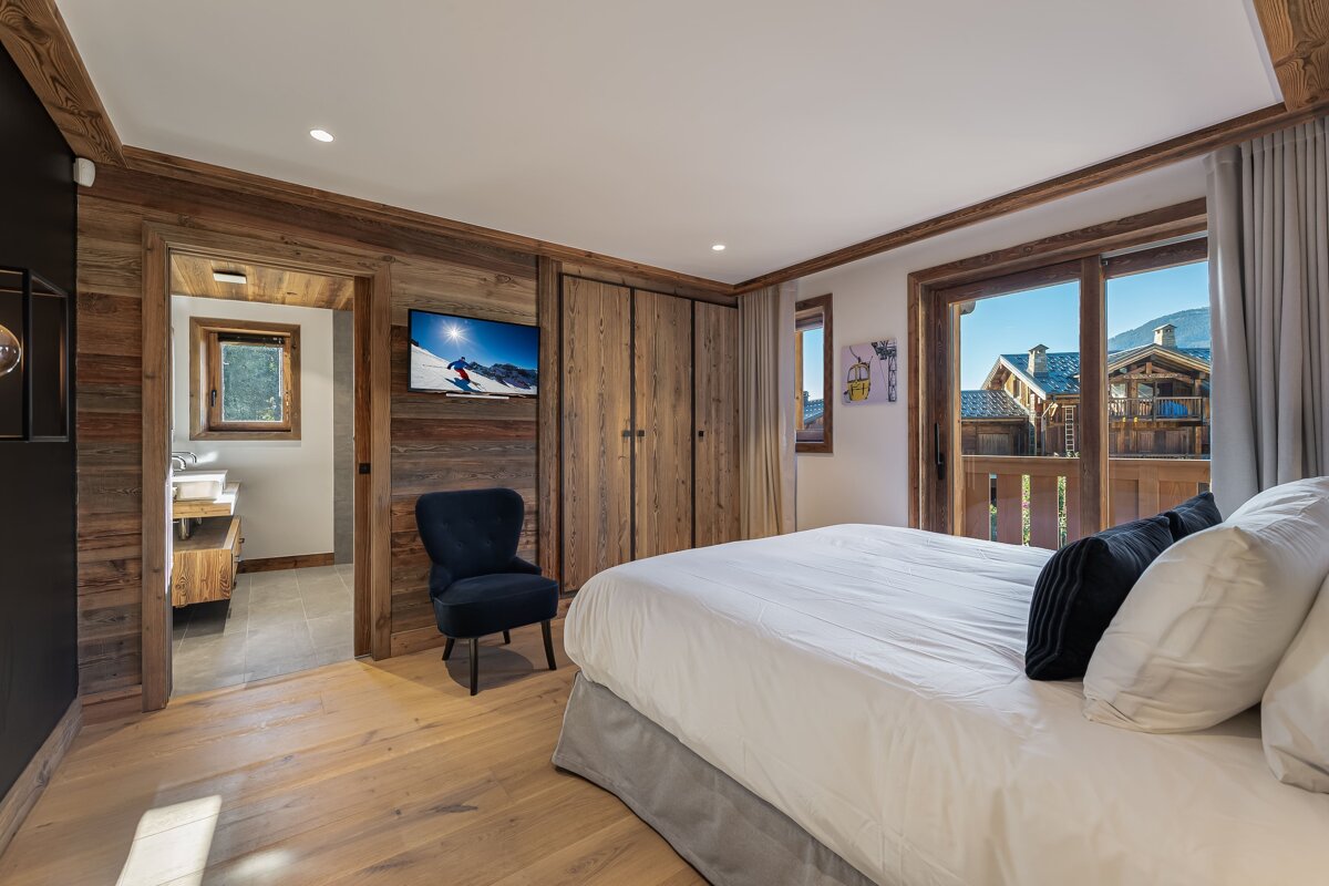 A cozy, rustic wooden bedroom featuring a large bed, TV, and a balcony with views of mountain chalets. An en-suite bathroom is visible.