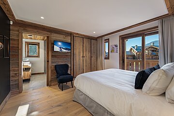 A cozy, rustic wooden bedroom featuring a large bed, TV, and a balcony with views of mountain chalets. An en-suite bathroom is visible.