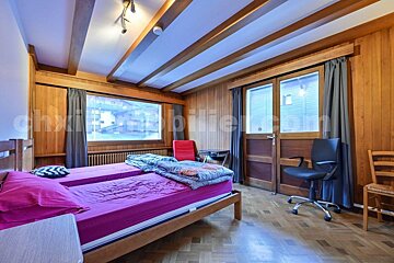 A cozy, wood-paneled room with twin beds, a large window overlooking buildings, and a door. Wooden ceiling beams add rustic charm.