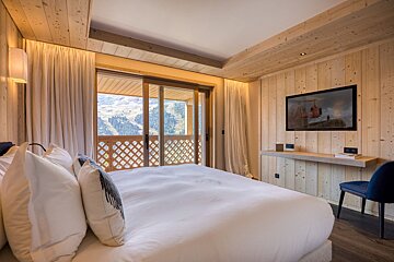 Cozy wooden chalet bedroom with a large white bed, wall-mounted TV, and sliding doors opening to scenic mountain views.