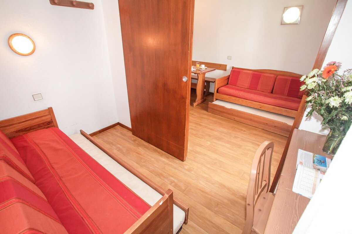 A small apartment interior showing two interconnected rooms with wooden floors. One has a pull-out bed, the other a red sofa-bed, dining set, and flowers on a desk.