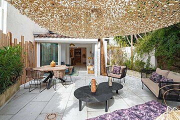 A modern patio features a camouflage shade, dining set, lounge furniture, and lush plants, leading into a stylish home.