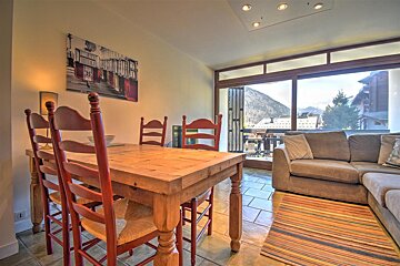 A cozy room features a wooden dining table with red chairs, a sofa, and large windows revealing scenic mountain village views.