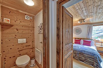 A wood-paneled toilet room is on the left, next to an open door revealing a rustic bedroom with a bed, red pillows, and a window view.