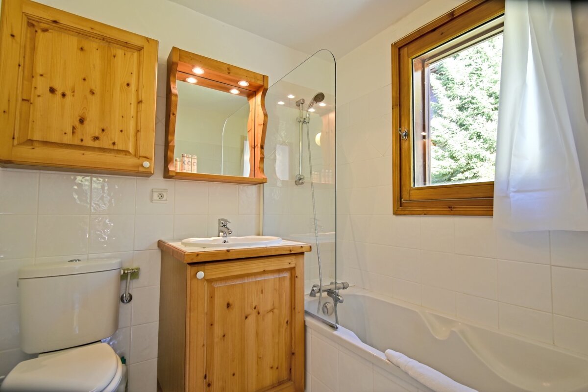 A rustic bathroom with wooden cabinets, a sink vanity, a toilet, and a bathtub with a glass screen next to a window.