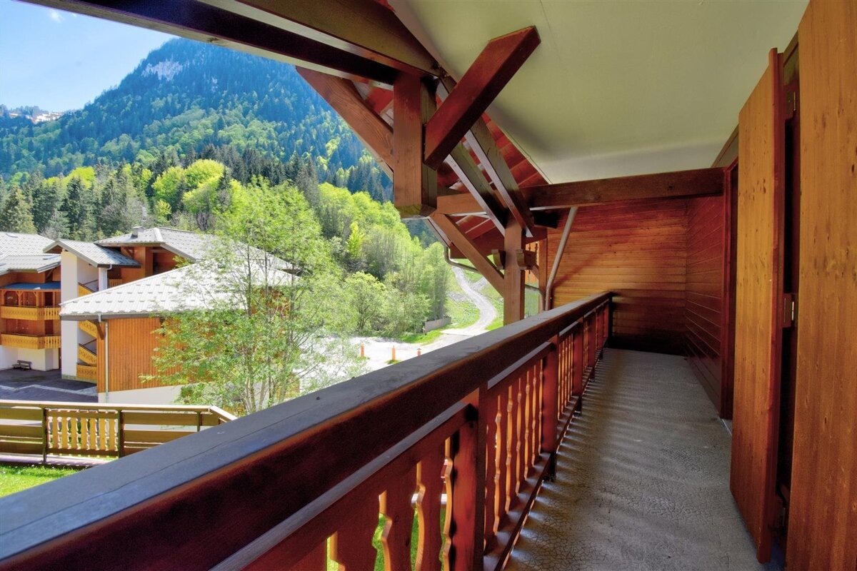 A wooden balcony offers a scenic view of lush green mountains, forests, and alpine buildings under a clear sky.