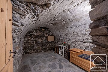 Rustic stone room with an arched, uneven ceiling, stone walls and floor, featuring a wooden bench, small table, and open door.