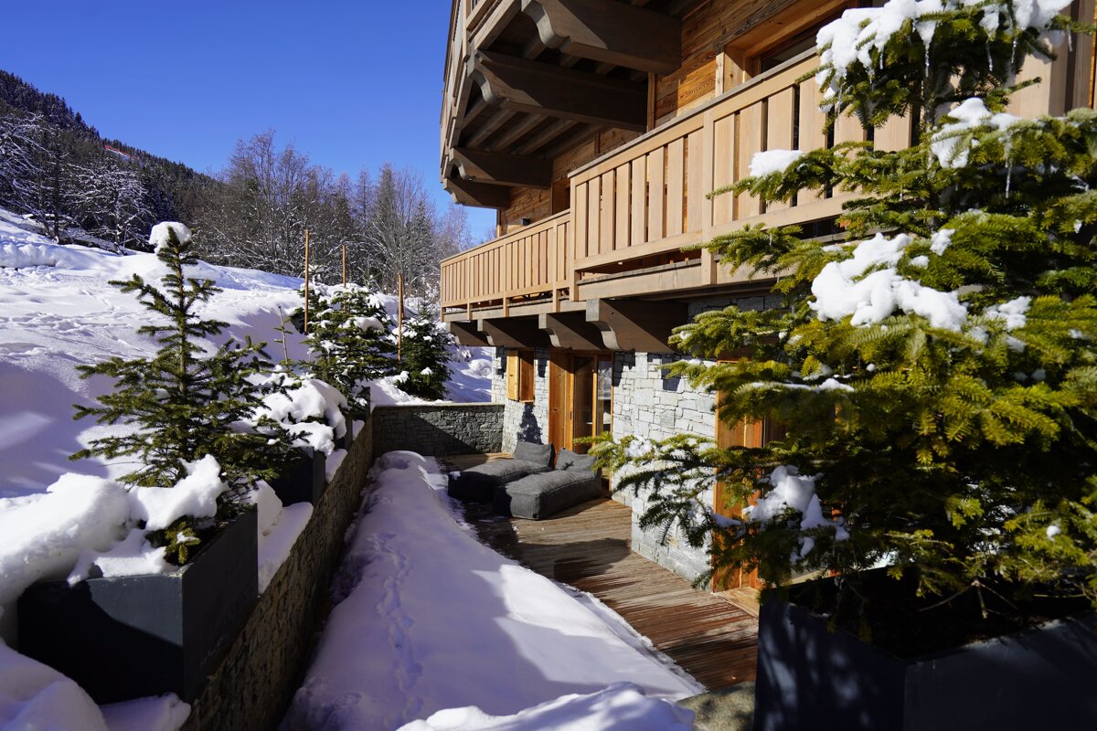 Sunny winter day at a wooden mountain chalet with snow-covered ground, evergreen trees, and a clear blue sky.