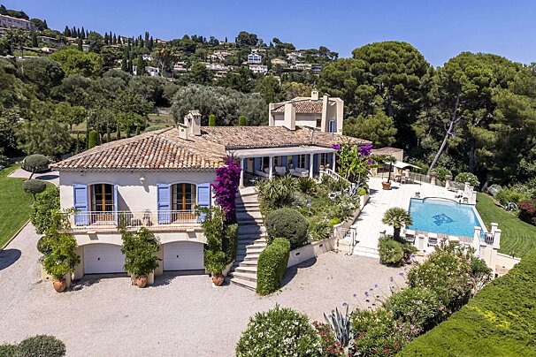 Sunny Mediterranean villa with terracotta roof, blue shutters, lush gardens, a private pool, and hillside views.