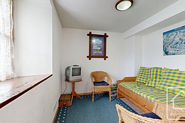 A cozy, rustic room with a wooden daybed, wicker chairs, old TV, and mountain-themed decor. A blue patterned rug covers the floor.