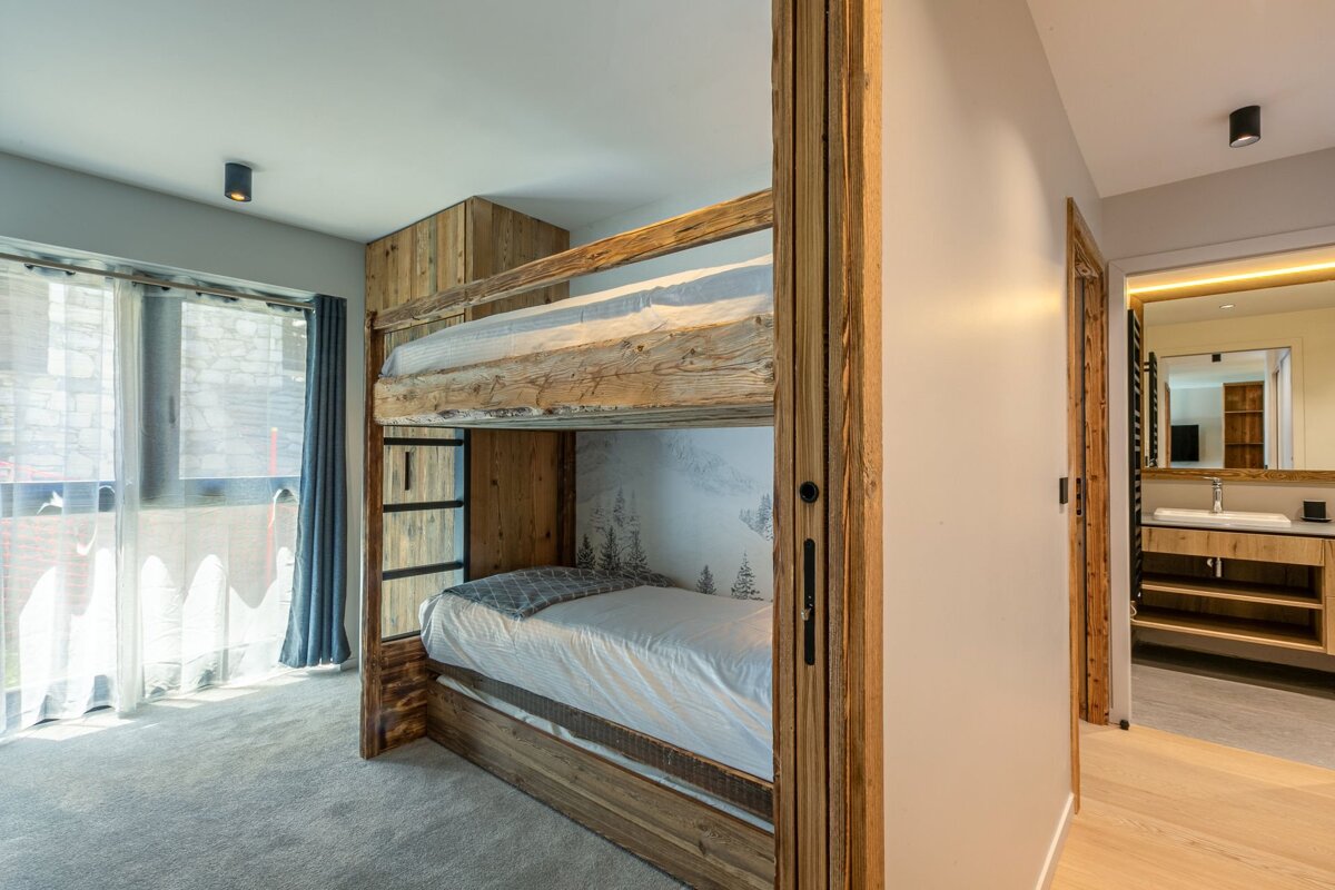 A stylish, rustic-modern bedroom features wooden bunk beds, a subtle mountain mural, a bright window, and an adjoining bathroom.