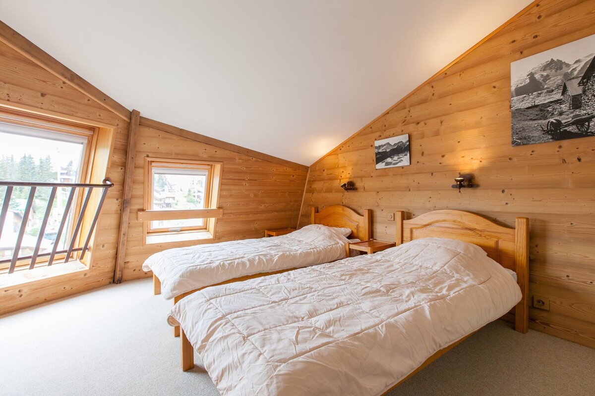 A cozy, wood-paneled chalet bedroom features a large bed, two windows with mountain views, and charming winter-themed decor.