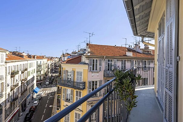 Balcony view of a sunny European city street with traditional buildings, terracotta roofs, and a clear blue sky. A plant adorns the railing.