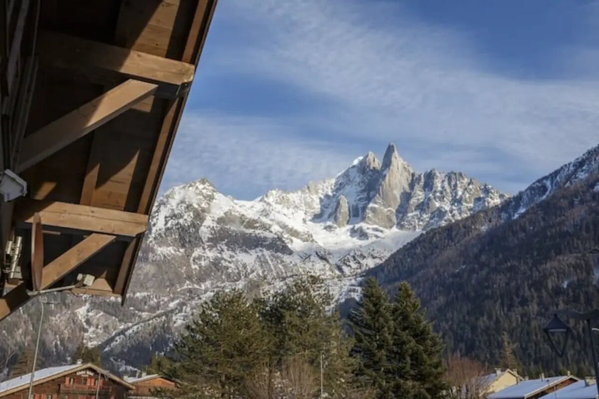 Majestic snow-covered mountain peaks rise behind evergreen trees, framed by the wooden eaves of an alpine chalet.