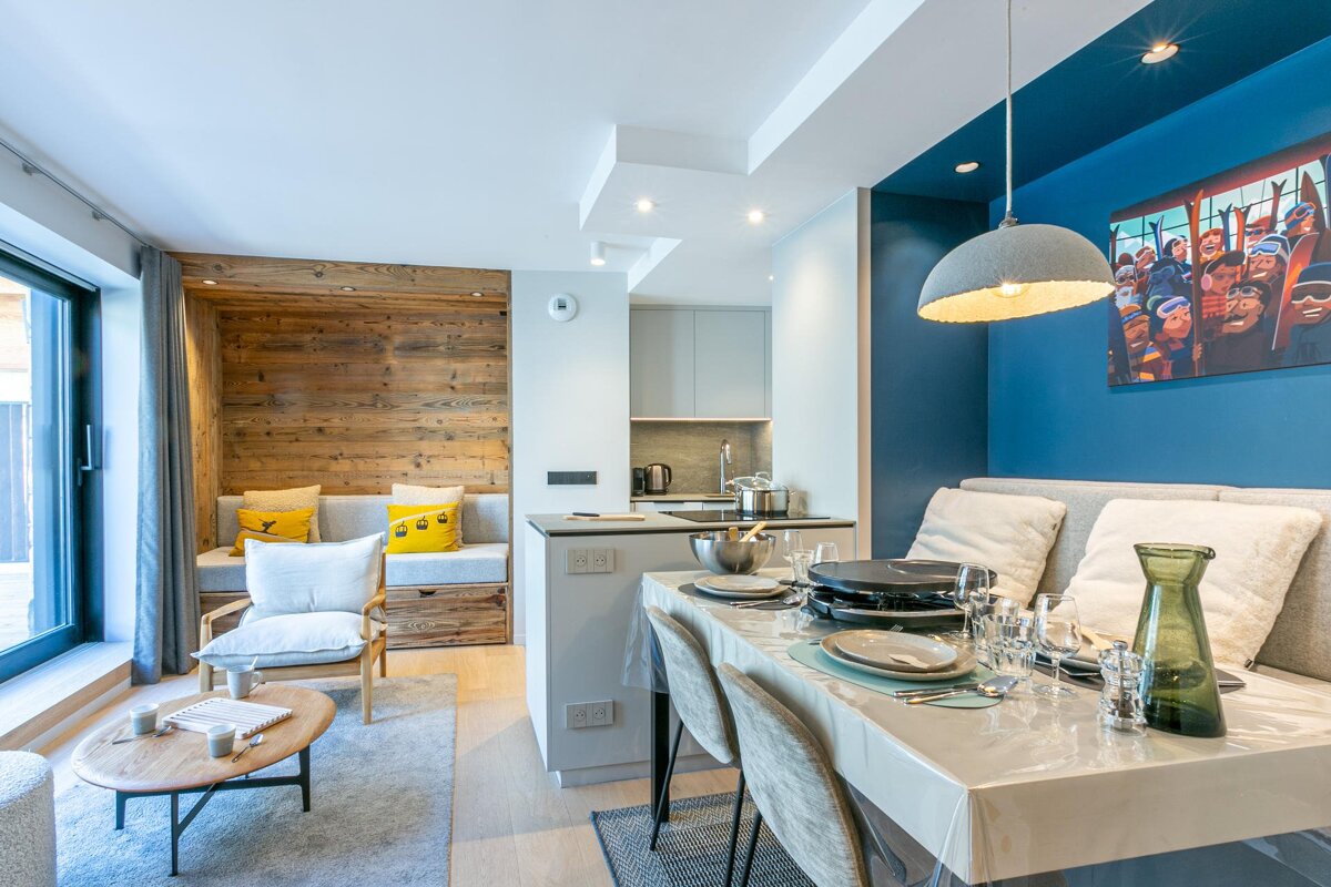 Modern open-plan living room, dining area with raclette, and kitchen. Features a cozy wooden nook and blue wall art.