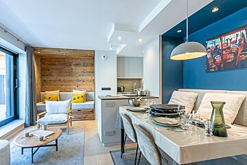 Modern open-plan living room, dining area with raclette, and kitchen. Features a cozy wooden nook and blue wall art.