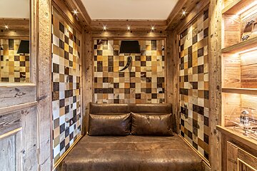 A rustic wood-paneled alcove with a brown leather bench, walls adorned with checkered hide squares, and a black wall lamp featuring a horn design.