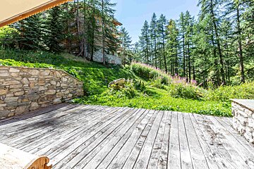 Wooden deck overlooking a lush green hillside with a stone wall, tall pine trees, and a mountain chalet under a clear sky.