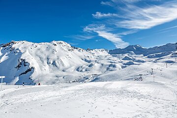 Expansive, sunny ski resort with snow-covered mountains, groomed slopes, and ski lifts under a clear blue sky. A few skiers are visible.