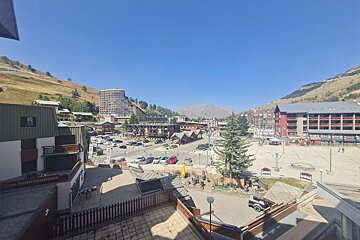 Sunny summer view of an alpine resort village with hotels, plazas, and surrounding grassy mountains under a clear blue sky.
