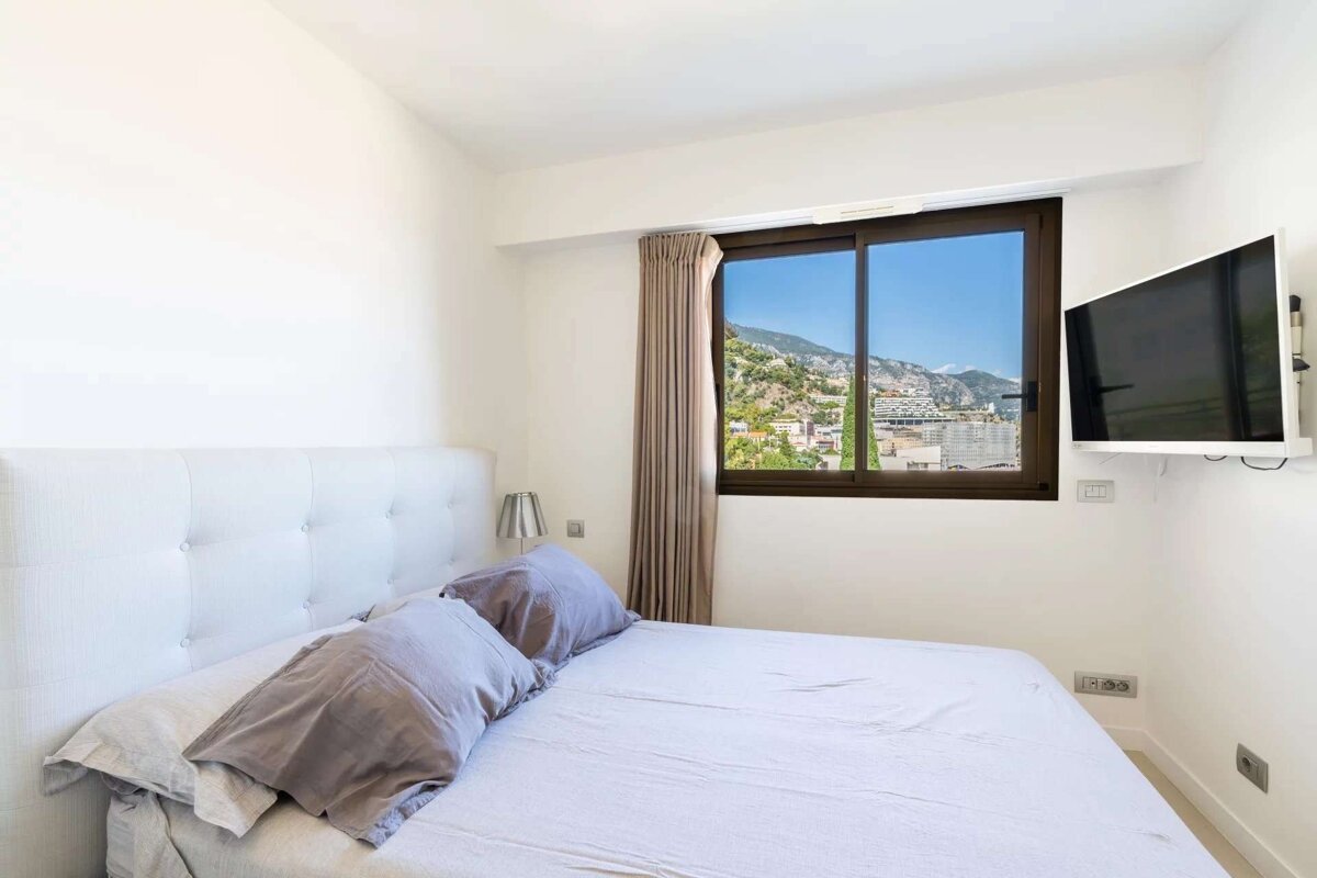 Bright bedroom with a white bed, TV, and a large window offering a scenic view of mountains and a city.