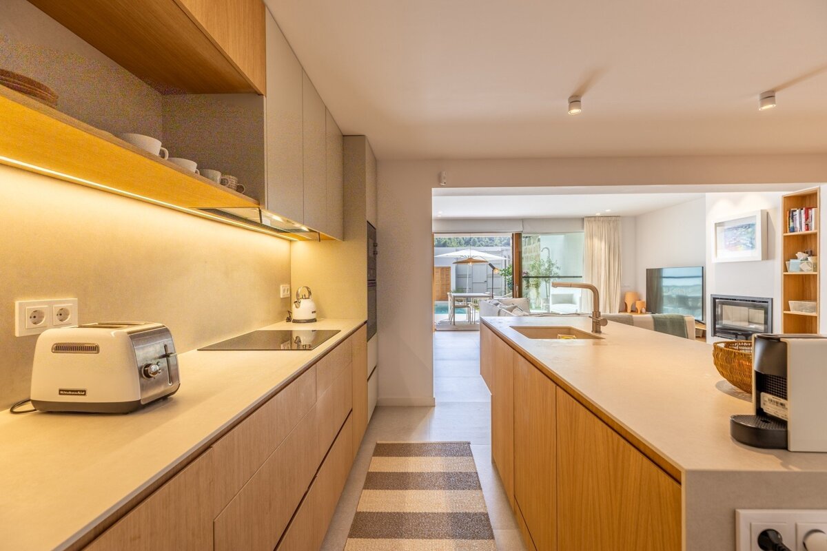 Bright, open-plan modern kitchen with light wood accents, light countertops, and warm lighting, connecting to a living room and outdoor pool.