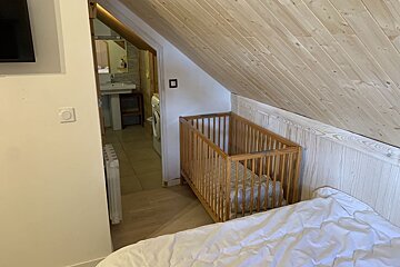 Cozy attic room with a sloped wooden ceiling, a baby crib under the eaves, and an open doorway leading to a small bathroom.