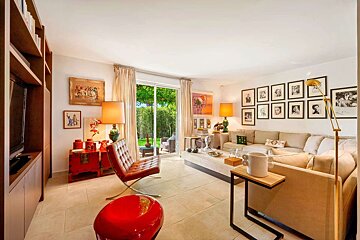 An eclectic living room with a large sofa, gallery wall, red accents, and a glass door opening to a lush garden.