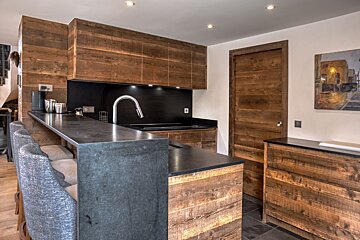 Modern kitchen with rustic wood cabinets, dark counters, and a breakfast bar with gray stools. Features a sleek faucet and a wall painting.