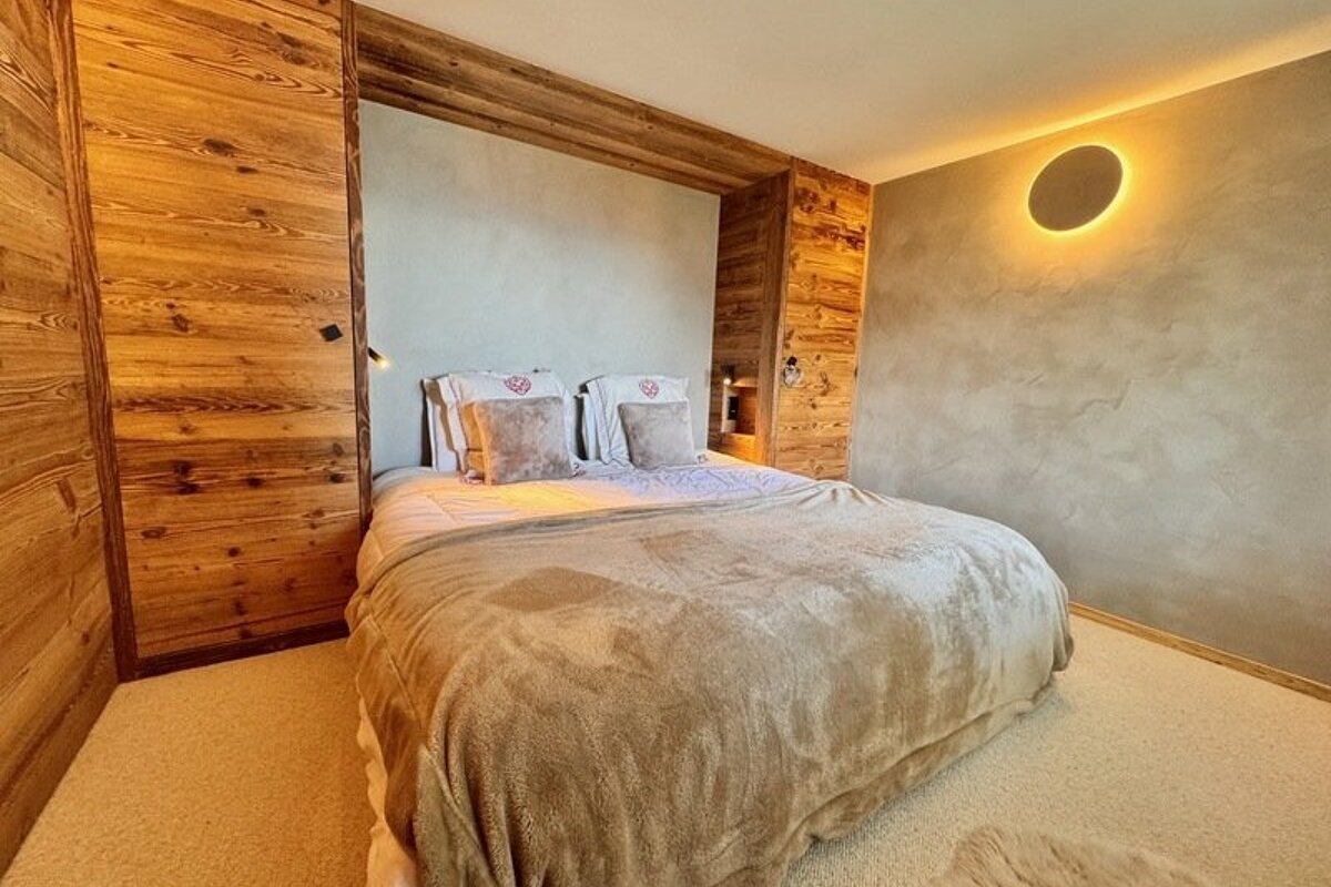 Cozy bedroom featuring a large bed with a plush beige blanket, warm wooden paneling, and a circular backlit wall light.