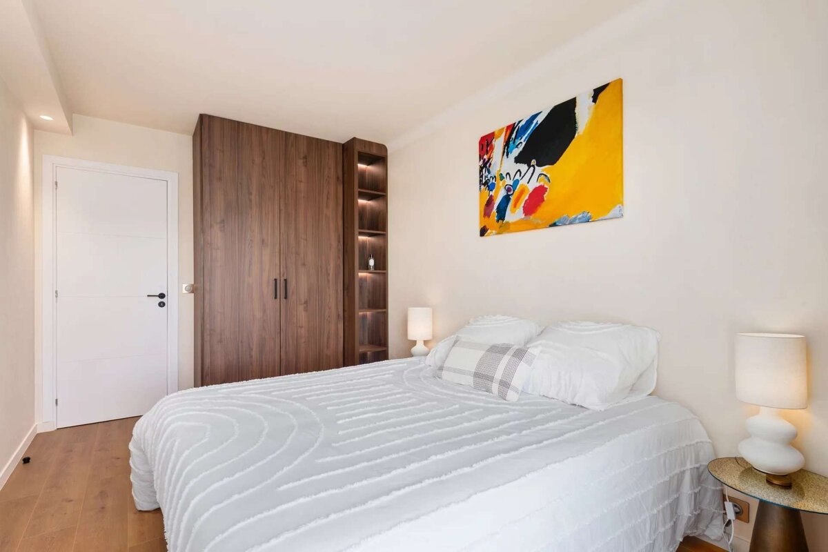 A bright, modern bedroom with a white bed, large wooden closet, and colorful abstract art. Features two white lamps, a white door, and hardwood flooring.