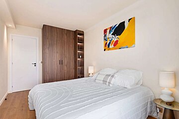 A bright, modern bedroom with a white bed, large wooden closet, and colorful abstract art. Features two white lamps, a white door, and hardwood flooring.