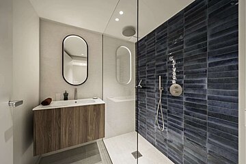 Modern bathroom with a wooden vanity, backlit oval mirror, and a walk-in shower featuring dark blue subway tiles and a glass partition.