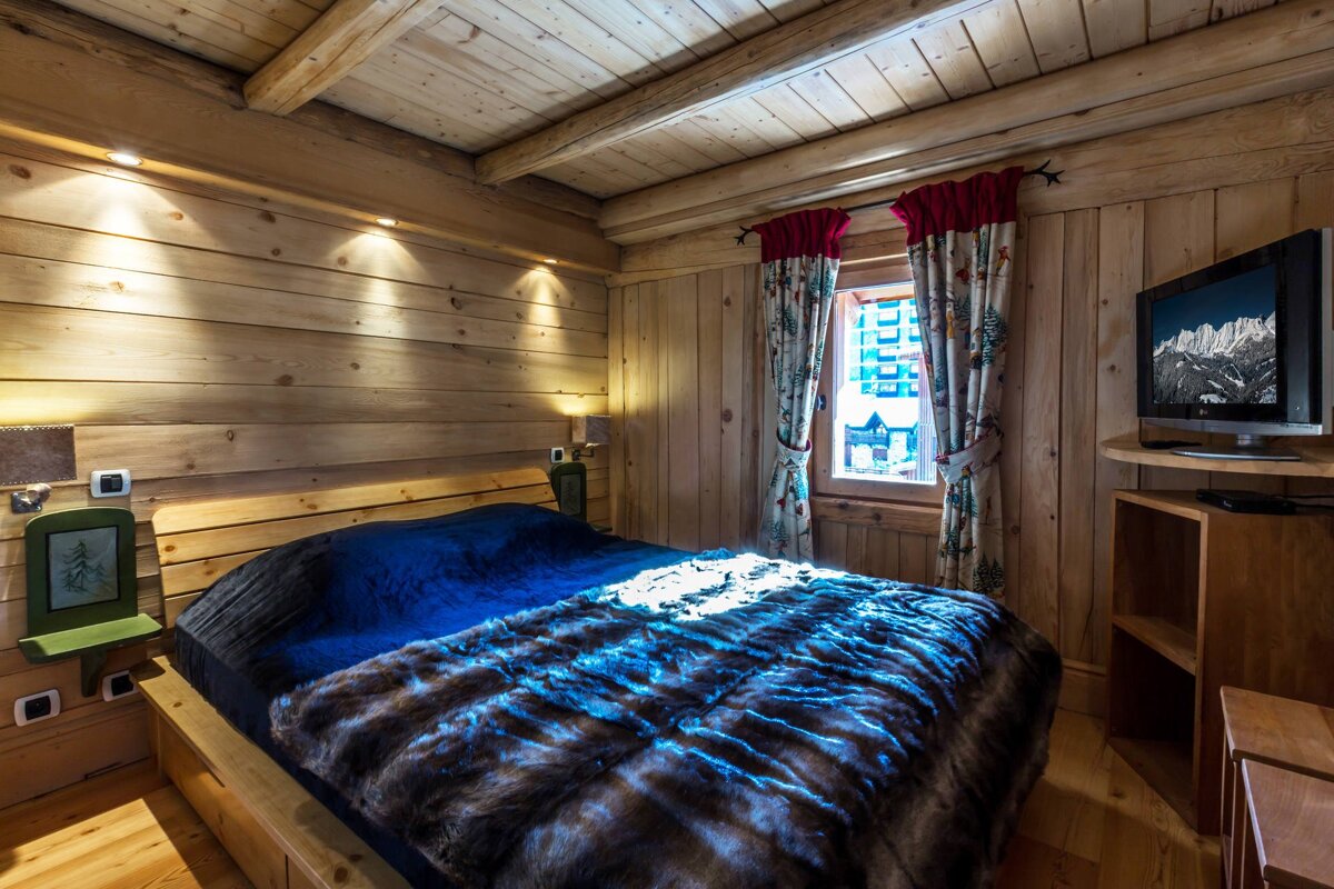 Cozy wooden cabin bedroom with a bed, blue and furry blanket, window with patterned curtains, and a TV showing snowy mountains.
