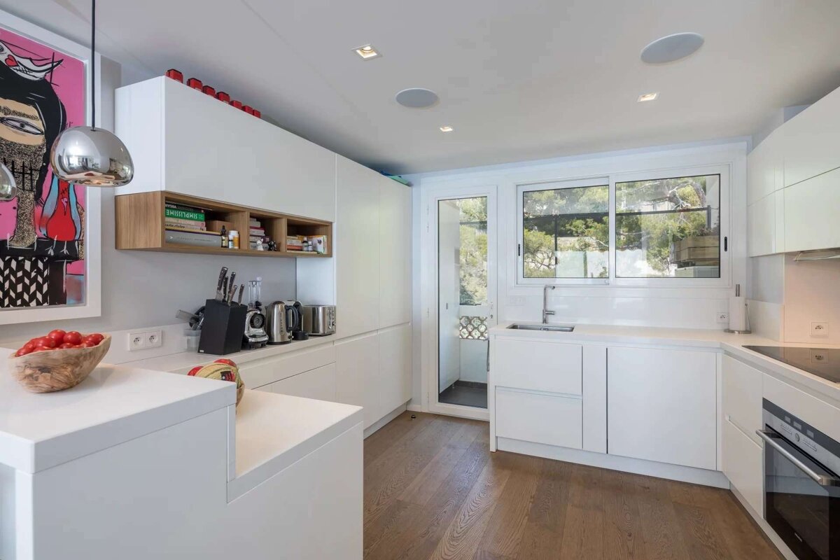 A bright, modern kitchen featuring white cabinets, wooden floors, and large windows revealing greenery. Artwork and a bowl of tomatoes add pops of color.