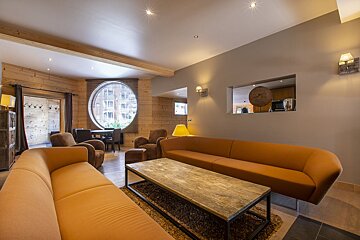 Cozy modern living room with curved orange sofas, a rustic wood coffee table, and a large circular window in a wood-paneled wall.
