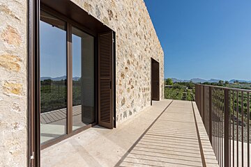A rustic stone building features a balcony with brown railings, glass doors, and shutters, offering views of a vineyard and mountains under a clear sky.