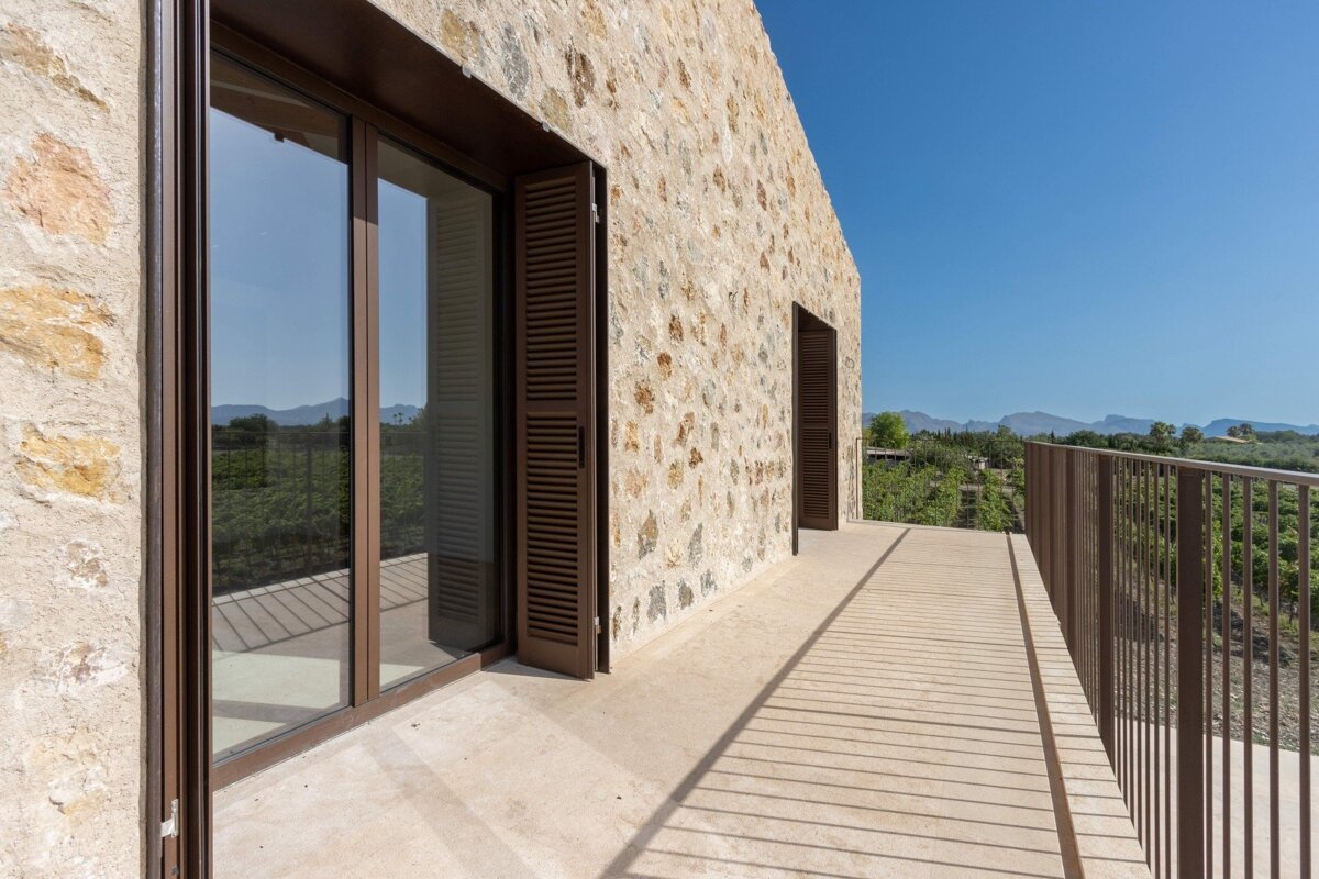 A stone-walled building with a balcony, glass doors, and open shutters, overlooking a green, mountainous landscape under a clear blue sky.