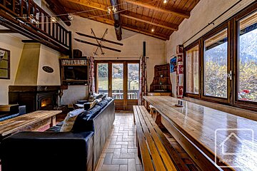 Cozy, rustic chalet living/dining room with wooden beams, fireplace, and large windows showcasing mountain views. Skis adorn the wall.