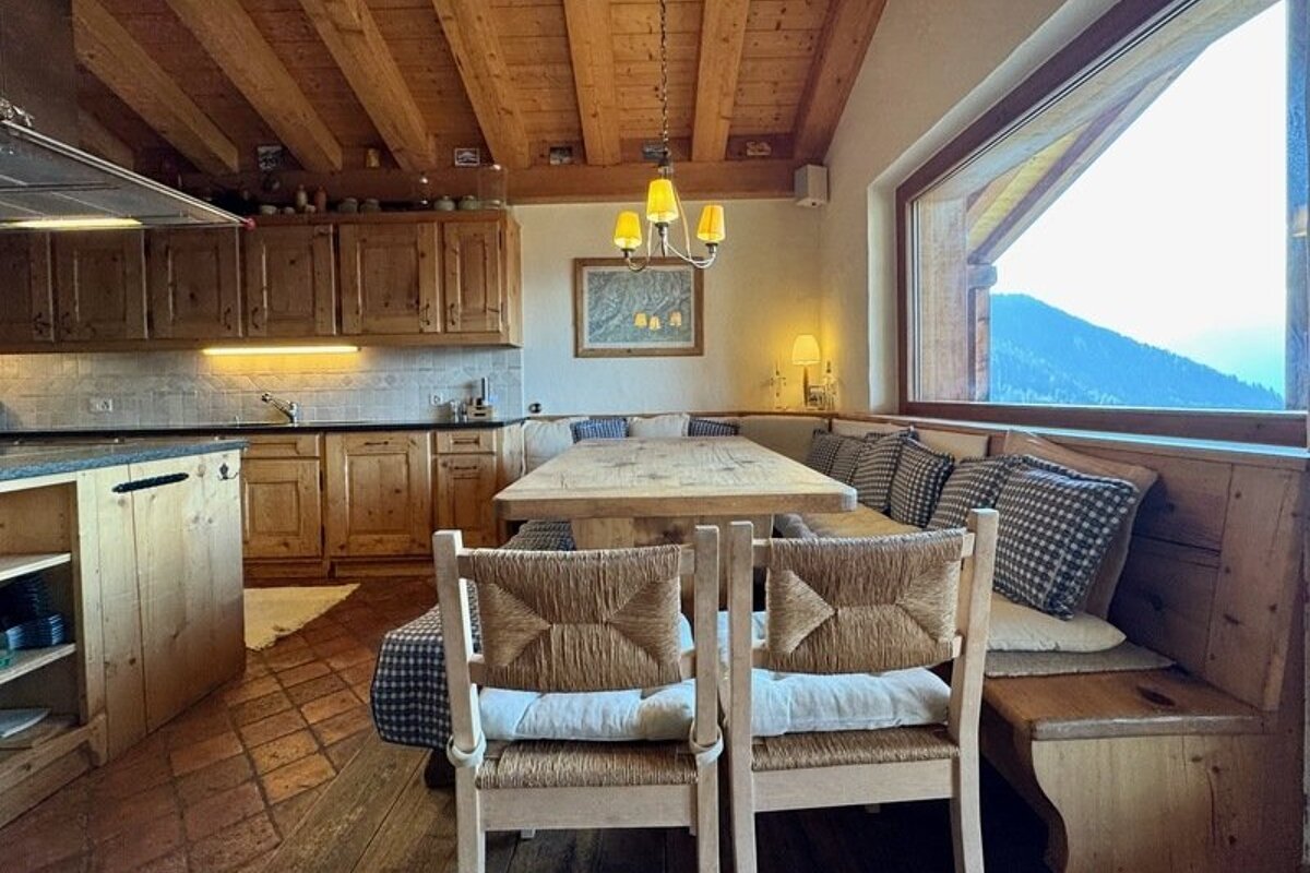 Cozy, rustic alpine kitchen and dining area with wooden furniture, warm lighting, and a large window with mountain views.