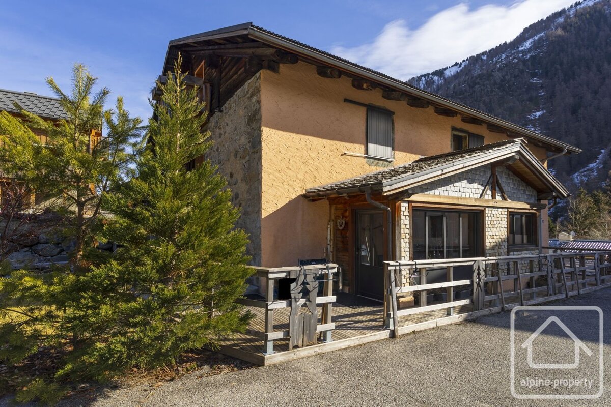 A rustic alpine chalet with stone and stucco walls, a wooden deck, and pine trees, set against a backdrop of sunny, snowy mountains.