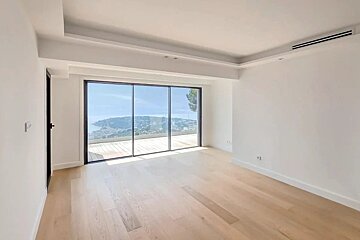 An empty modern room with light wood floors, white walls, and large sliding glass doors offering a beautiful coastal view.