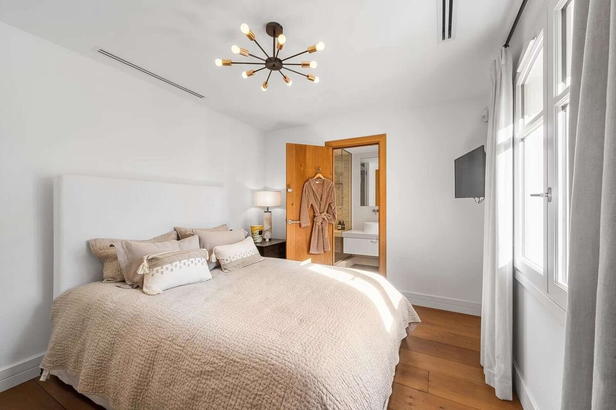 A bright bedroom with a large bed, neutral quilt, modern ceiling light, wood floor, and an open bathroom door with a robe hanging.