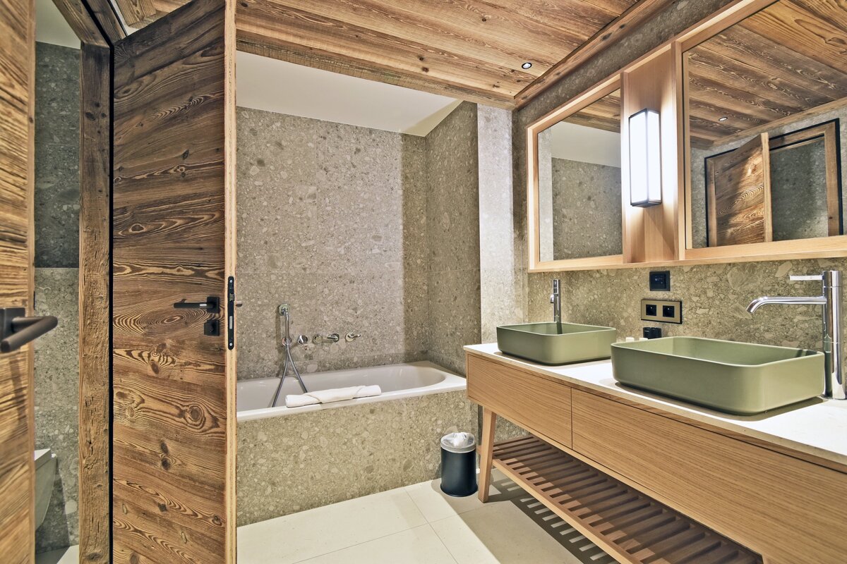 Chic modern bathroom with a wooden door, ceiling, and stone walls. Features a bathtub and a double vanity with sage green vessel sinks and mirrors.