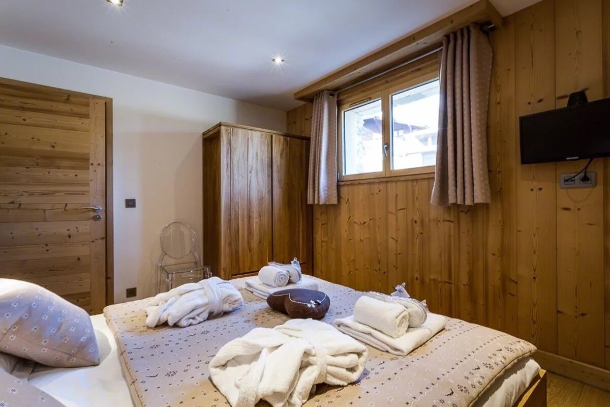 A cozy, wood-paneled bedroom features a bed neatly prepared with folded towels and white robes, a wooden wardrobe, and a window.