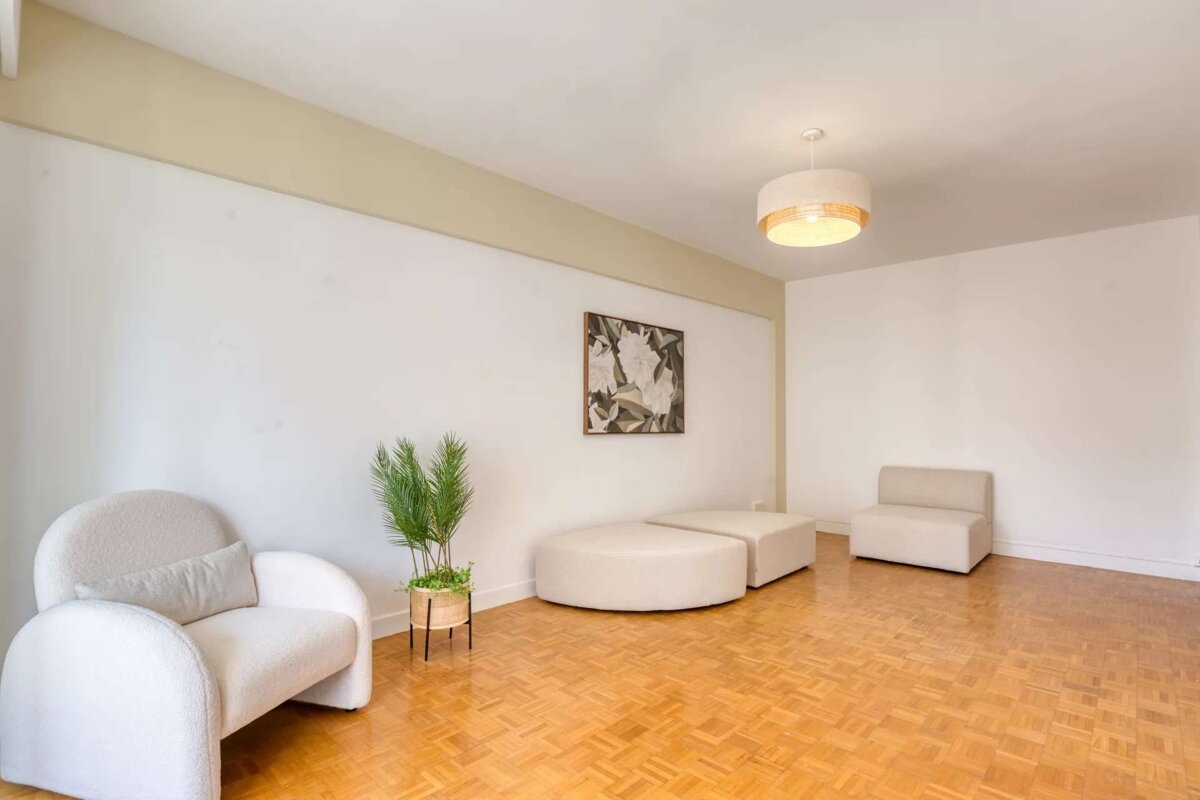 A bright, minimalist room featuring white walls, light parquet floors, cream modular seating, a stylish armchair, green plant, and abstract wall art.