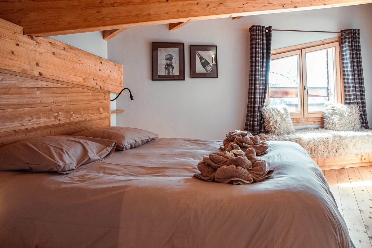Cozy cabin bedroom featuring a wooden headboard, bed with folded towels, and a sunny window seat with fluffy cushions.