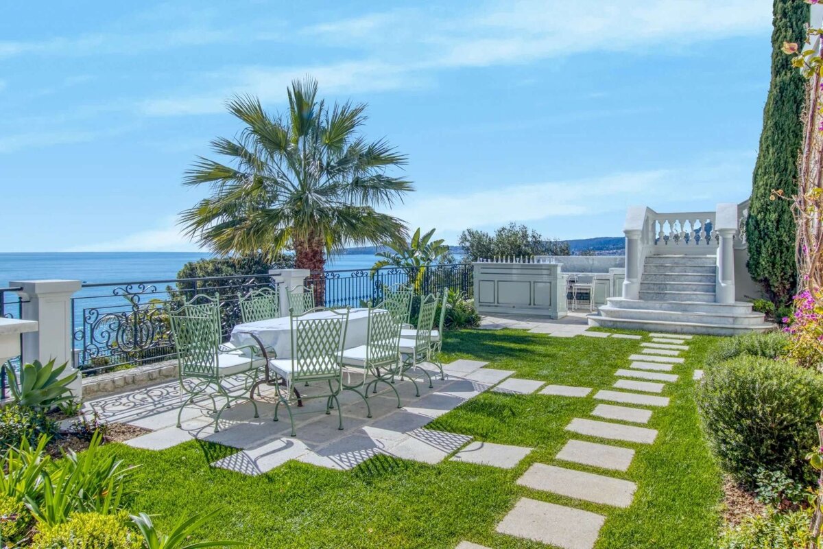 A luxurious outdoor patio overlooks a blue ocean. It features a dining set on a stone terrace, a green lawn with stepping stones, a palm tree, and elegant white stairs under a clear sky.