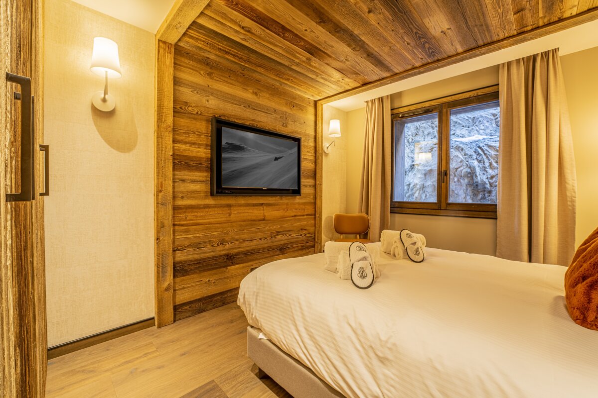 Cozy chalet bedroom featuring warm wood paneling, a wall-mounted TV, and a bed with towels and slippers.